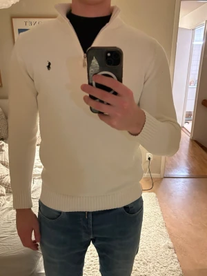 Vit halvzip-tröja från Ralph Lauren - Klassisk vit stickad tröja från Ralph Lauren med halv dragkedja och broderad logga på bröstet. Tröjan har ribbade muddar och krage, perfekt för en clean och stilren look. Passar lika bra till jeans som till chinos.