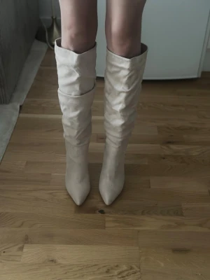 Ljusa slouchy knähöga stövlar - Säljer ett par ljusa, slouchy knähöga stövlar med spetsig tå och bred klack. Stövlarna har en mjuk mockaliknande yta och en avslappnad passform som ger en trendig look. Perfekta att styla till kjol eller jeans för en cool vibe.