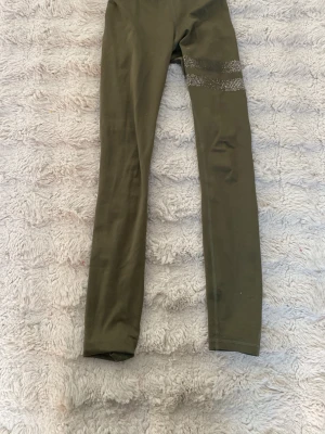 Olivgröna tights från Stronger - Snygga olivgröna tights från Stronger med dekorativa silverfärgade detaljer runt ena låret. Tightsen har elastisk midja med logotyp och är tillverkade i ett stretchigt material som passar perfekt för träning eller gym.
