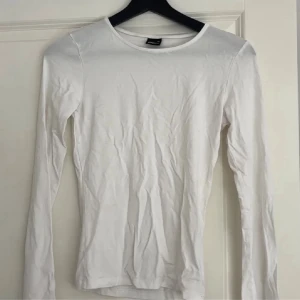Vit långärmad topp från Gina Tricot - En basic vit långärmad topp från Gina Tricot med rund halsringning. Tunn och mjuk bomullskänsla, perfekt att styla lager på lager eller till jeans. Enkel och stilren design som funkar till många outfits.
