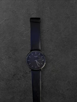 Svart klocka från Regal med läderband - Stilren svart klocka från Regal med rund urtavla och minimalistisk design. Klockan har ett svart läderband och silverfärgade markeringar på urtavlan. Perfekt accessoar för en clean och modern look.