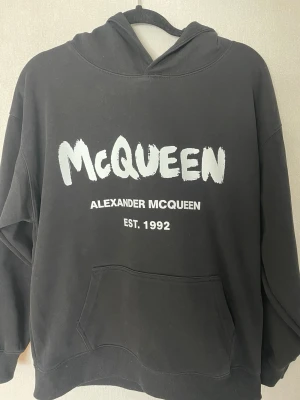 Alexsander McQueen hoodie - Svart hoodie från Alexander McQueen med stor vit logga och texten 'EST. 1992' på bröstet. Klassisk känguruficka framtill och huva. Oversized passform och ribbade muddar. Perfekt för en streetwear-look.
