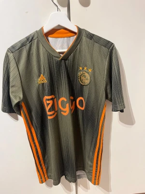 Ajax matchtröja adidas grön/orange - Säljer en Ajax bortamatchtröja från säsongen 21/22. Tröjan har orange detaljer, klubbmärke och sponsortryck framtill samt tre stjärnor ovanför loggan. Tillverkad i lätt och ventilerande polyester, perfekt för fotbollsträning eller en skön sommardag.