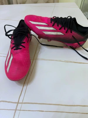 Elite Adidas rosa fotbollsskor med dobbar - Säljer ett par snygga fotbollsskor från Adidas i en stark rosa färg med svarta detaljer och vita stripes på sidorna. Skorna har snörning och dobbar för bra grepp på planen. Ovandelen är i syntetmaterial och sulan är designad för fart och kontroll.
