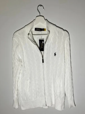 Vit kabelstickad hel ziptröja Polo Ralph Lauren🙌 - Kabelstickad vit tröja från Polo Ralph Lauren med dragkedja framtill och ribbade muddar. Klassisk broderad logga på bröstet i marinblått. Tröjan har hög krage och två fickor framtill. Perfekt för en clean och stilren look.