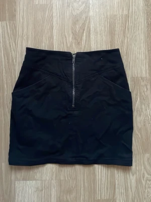 Svart kortkjol med dragkedja - Snygg svart kortkjol med markerad midja och en tydlig dragkedja framtill. Kjolen har en enkel, stilren design och är lätt att matcha med olika toppar. Perfekt för dig som gillar en clean och modern look.