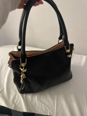 Svart handväska med gulddetaljer - Snygg svart handväska i slätt konstläder med två rejäla handtag och guldfärgade metalldetaljer. Väskan har en rosa kant Baksidan har en liten ficka med dragkedja. 