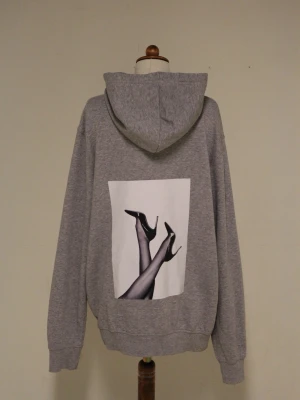 Grå hoodie från Reuse med tryck - Snygg grå hoodie från Reuse med huva och ficka. På bröstet finns en liten svart logga och på ryggen ett stort tryck med ben i högklackat. Tillverkad i mjuk bomullsblandning, perfekt för en avslappnad streetwear-look.