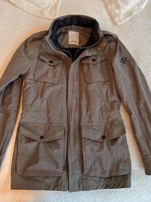 J.Lindeberg jacka - Snygg grå field jacka från J.Lindeberg med fyra stora fickor framtill och patch på ärmen. Jackan har hög krage, dragkedja och tryckknappar. Tillverkad i lätt och slitstarkt material, perfekt för blåsiga dagar.