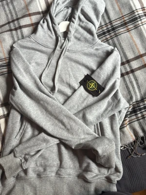 Grå hoodie från Stone Island - Snygg grå hoodie från Stone Island med klassisk huva och dragsko. Tröjan har den ikoniska svarta patchen med gul logga på ärmen. Tillverkad i mjuk bomullsmix och har ribbade muddar vid ärmslut och nederkant. Perfekt för en avslappnad streetwear-look.