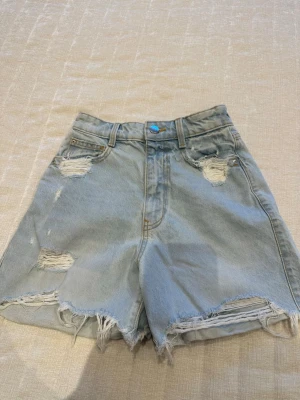 Ljusa denimshorts från ZARA - Snygga ljusblå denimshorts från ZARA med slitna detaljer och fransig kant. Klassisk femficksmodell med hög midja och knappstängning. Perfekta för en avslappnad och trendig look.