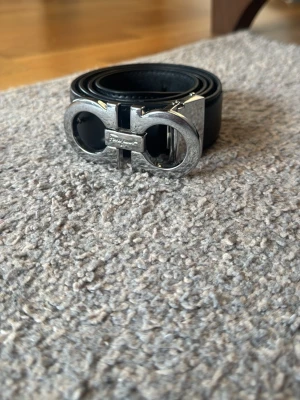 Svart skärp från Salvatore Ferragamo - Stilrent svart skärp från Salvatore Ferragamo med ikoniskt dubbel-Gancini-spänne i silverfärgad metall. Skärpet är gjort i slätt skinn och passar perfekt för att lyfta din outfit med en lyxig touch.