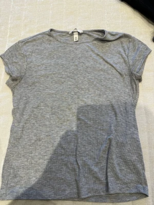 Grå ribbad t-shirt från H&M - En enkel och stilren grå ribbad t-shirt från H&M. T-shirten har korta ärmar, rund halsringning och är tillverkad i mjuk bomull. Perfekt basplagg att matcha med allt i garderoben.