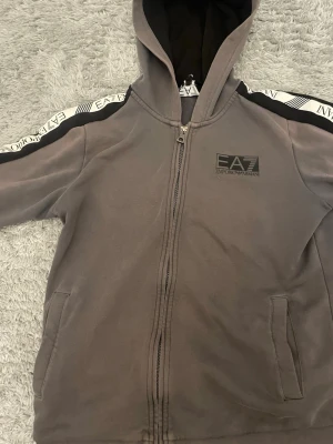 Grå hoodie från EA7 Emporio Armani - Snygg grå hoodie från EA7 Emporio Armani med dragkedja framtill och svart logga på bröstet. Svarta detaljer på axlarna och vita band med EA7-tryck längs ärmarna. Två fickor fram och huva med svart innerfoder. lite små defekter på vissa ställen men det är inget man ser eller tänker på.