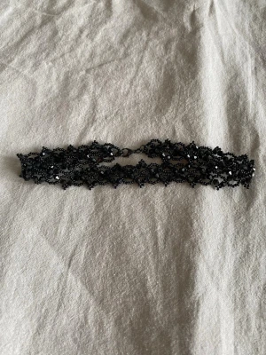 Handgjord svart pärlchoker med blommönster - Säljer en svart choker i pärlor med ett detaljerat blommönster. Halsbandet har små glittrande pärlor som ger en cool och edgy vibe. Perfekt för att lyfta vilken outfit som helst med en unik touch. Stängs med en liten metallhake bak.