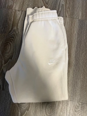 Beige mjukisbyxor från Nike - Snygga beige mjukisbyxor från Nike med broderad logga på benet. Byxorna har resår i midjan och vid bensluten för en skön passform. Perfekta för chill dagar eller när du vill ha en avslappnad stil. Tillverkade i mjukt material som känns bekvämt hela dagen.Skriv för fler frågor och bilder dress finns 