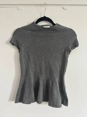 Grå finstickad peplumtopp med kort ärm - En enkel och stilren grå finstickad topp med korta ärmar. Toppen har en lätt utsvängd passform nedtill och är perfekt för att matcha med jeans eller kjol. Materialet känns mjukt och stretchigt.