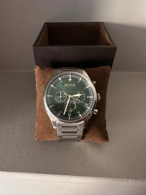 Silverfärgad BOSS klocka med grön urtavla - Stilren klocka från BOSS med silverfärgad länk i metall och grön urtavla. Klockan har kronograf-funktion, tre mindre urtavlor och tydliga vita markeringar. Klassisk design som passar till många olika looks.