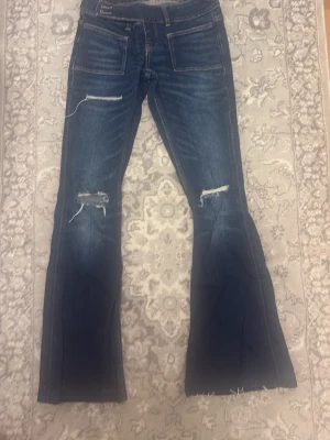 Blå bootcut jeans från Gina Tricot - Säljer ett par blå bootcut jeans från Gina Tricot i storlek XS. Jeansen har slitna detaljer vid knäna, råa kanter nertill och markerade sömmar. Klassisk femficksmodell med låg midja och stretchigt denimtyg.