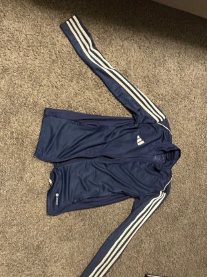 Marinblå adidas träningsjacka XS - Snygg marinblå träningsjacka från adidas med klassiska vita ränder längs ärmarna och dragkedja framtill. Jackan har hög krage och är tillverkad i ett lätt, sportigt material som passar perfekt till träning eller chill. Ikonisk adidas-logga på bröstet.