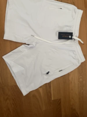 Vita shorts från Polo Ralph Lauren - Snygga vita shorts från Polo Ralph Lauren med dragsko i midjan och klassisk logga broderad på benet. Shortsen har fickor både fram och bak och är tillverkade i ett mjukt bomullsmaterial. Perfekta för sommardagar och enkel att matcha.