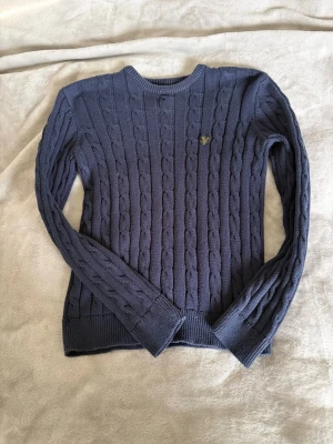 Mörkblå kabelstickad tröja Lyle & Scott - Kabelstickad mörkblå tröja från Lyle & Scott med rund halsringning och ribbade muddar. Tröjan har det klassiska Lyle & Scott-logotypen broderad på bröstet och är tillverkad i mjukt material. Perfekt för dig som gillar stilren och tidlös design.