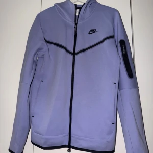 Ljuslila Nike hoodie med dragkedja - Nike hoodie i ljuslila med hel dragkedja framtill och svart Nike-logga på bröstet. Hoodien har svarta detaljer, fickor på sidorna och en extra ficka med dragkedja på ena ärmen. Tillverkad i ett mjukt och stretchigt material. Perfekt för en sportig och avslappnad stil.