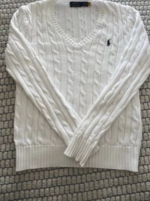 Vit kabelstickad tröja Polo Ralph Lauren - Kabelstickad vit tröja från Polo Ralph Lauren med V-ringning och klassisk marinblå logga broderad på bröstet. Tröjan har långa ärmar, ribbade muddar och är gjord i mjukt material som passar perfekt till lager-på-lager-stil.