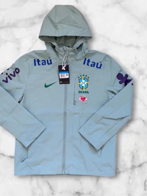 Nike Brasil vindjacka med huva - Ljusgrön vindjacka från Nike med Brasiliens landslagslogga och flera sponsortryck i blått och lila. Jackan har huva, hel dragkedja och långa ärmar. Perfekt för dig som gillar sportig stil och vill sticka ut med landslagsvibe.