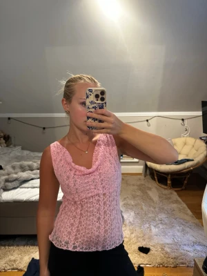 Rosa spetslinne med draperad hals - Säljer ett ljusrosa ärmlöst spetslinne med draperad halsringning. Toppen har ett feminint och romantiskt uttryck med transparent spetsmaterial och en lös passform. Perfekt att styla med jeans eller kjol för en trendig look.