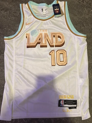 Cleveland Cavaliers basketlinne Nike - Snyggt vitt basketlinne från Nike med Cleveland Cavaliers 'The Land' tryck och nummer 10 i guldbrunt. Linnet har ljusblå och guldbruna detaljer runt hals och ärmhål. Tillverkat i lätt och ventilerande meshmaterial, perfekt för basket eller streetwear.