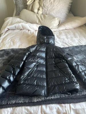 Svart dunjacka från Moncler - Snygg svart dunjacka från Moncler med glansig finish, huva och två stora dragkedjefickor framtill. Jackan har en ficka med Moncler-logga på ärmen och quiltad design som ger en modern look. Perfekt för kalla vinterdagar.