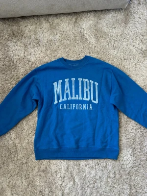 Blå sweatshirt med Malibu-tryck - Cool blå sweatshirt med stort vitt tryck 'MALIBU CALIFORNIA' på bröstet. Klassisk rund halsringning och långa ärmar. Perfekt för en avslappnad och trendig look. Mjuk och skön bomullsmix. Passar från xxs - s 