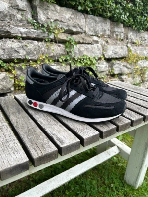 Svarta Adidas sneakers med silvriga ränder - Snygga svarta Adidas sneakers med klassiska silvriga ränder på sidorna och vita sulor. Skorna har svarta skosnören och detaljer i rött på sulan. Ovandelen är i mesh och mocka för en sportig look.