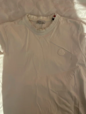 Vit Moncler t-shirt med logga - Säljer en stilren vit t-shirt från Moncler med rund hals och diskret Moncler-logga i vitt på bröstet. T-shirten är kortärmad och gjord i mjuk bomull som känns skön mot huden. Perfekt för dig som gillar minimalistisk design och exklusiva märken. dubbel krage, smått använd. FINNS I SVART