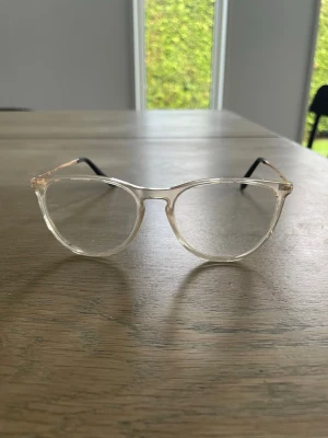 Ray-Ban transparenta glasögon - Säljer ett par stilrena glasögon från Ray-Ban med transparent båge och tunna guldfärgade skalmar. Skalmarna har svarta ändar för extra komfort. Perfekt accessoar för en trendig look.