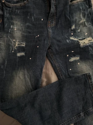 Mörkblå slitna jeans med färgstänk - Säljer ett par mörkblå jeans med slitningar och hål på benen samt vita färgstänk för en cool, trendig look. Jeansen har klassisk femficksmodell och raka ben. Perfekta för dig som gillar streetstyle och vill sticka ut lite extra.