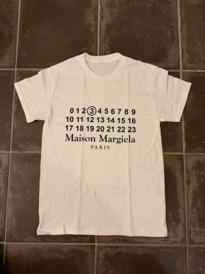 Maison Margiela vit t-shirt med tryck - Snygg vit t-shirt från Maison Margiela med det ikoniska numrerade trycket på bröstet och texten 'Maison Margiela PARIS' i svart. Klassisk rund hals och korta ärmar. Perfekt för dig som gillar minimalistisk och trendig design. Är ca 182 och väger 66