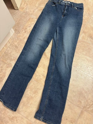 Blå bootcut jeans med broderade fickor - Säljer ett par klassiska blå bootcut jeans med snyggt broderade bakfickor och hög midja. Jeansen har en lätt tvättad look och är gjorda i mjukt denimtyg. Perfekta för en avslappnad och trendig stil.
