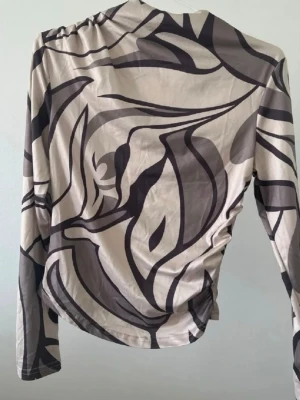 Beige och brun mönstrad långärmad topp - Trendig långärmad topp i beige med abstrakt brunt mönster. Tunn och stretchig med figurnära passform och asymmetrisk drapering på sidan. Perfekt för dig som gillar unika prints och vill sticka ut.