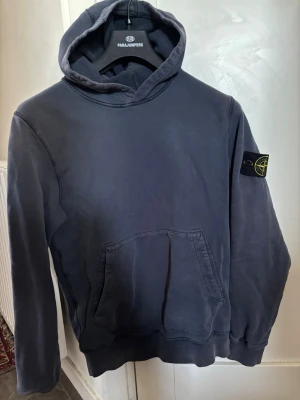 Mörkblå hoodie från Stone Island storlek S - Snygg mörkblå hoodie från Stone Island med klassisk patch på vänster ärm. Hoodien har en stor känguruficka framtill och ribbade muddar vid ärmslut och nederkant. Tillverkad i mjukt bomullsmaterial och har en avslappnad passform med huva. Storlek small. Inga fel på tröjan, knappt använd.
