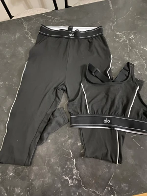 Svart set från Alo med vita detaljer S - Snyggt svart set från Alo med sport-bh och tights. Setet har vita kontrastsömmar längs sidorna och bred resår i midjan och under bysten med Alo-logga. Materialet är stretchigt och mjukt, perfekt för en aktiv livsstil.
