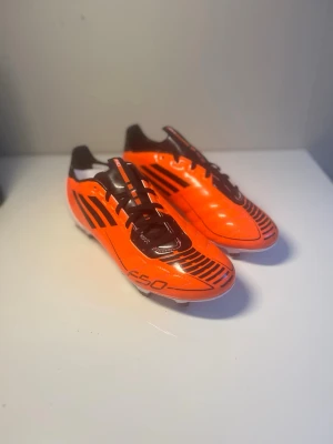 Adidas F50 orange fotbollsskor FG (äldre modell) - Snygga fotbollsskor från Adidas F50-serien i en skarp orange färg med svarta detaljer och vita sulor. Skorna har klassisk snörning och är designade för att ge bra grepp på planen. Perfekta för dig som vill sticka ut på matchen.
