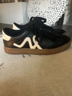 Svarta sneakers med vit detalj  - Snygga svarta sneakers från Kappa med vit vågig logga på sidan och vit häl. Skorna har svarta skosnören och en brun gummisula. Tillverkade i läderimitation och textil för en klassisk streetlook.