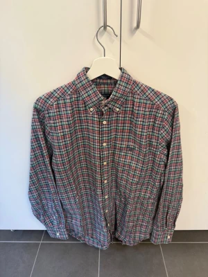 Rutig skjorta från Gant i bomull - Klassisk långärmad skjorta från Gant med rött, grönt, blått och vitt rutmönster. Skjortan har knappar framtill, button-down krage och en liten bröstficka med Gant-logga. Tillverkad i mjuk bomull, perfekt för en avslappnad men snygg stil.