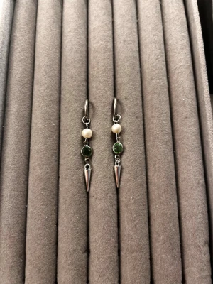 Silvriga örhängen med pärlor och grön sten - Säljer ett par snygga örhängen i silverfärg med vita pärlor, en mörkgrön rund sten och en spetsig metallisk detalj längst ner. Örhängena har en modern och edgy look som sticker ut. Perfekta för dig som gillar unika accessoarer.