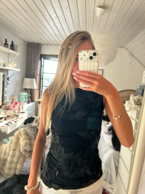 Svart blommig ärmlös topp - Snygg svart ärmlös topp med stora blommiga broderier. Toppen har en tight passform och är perfekt för dig som vill sticka ut lite extra. Materialet känns mjukt och stretchigt, och den höga halsen ger en clean look.