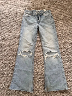 Ljusblå slitna bootcut jeans - Säljer ett par ljusblå bootcut jeans med stora slitningar över knäna och råa kanter nertill. Jeansen har klassisk femficksmodell och är tillverkade i denim. Perfekta för en avslappnad och trendig look.                       Tar emot bud!