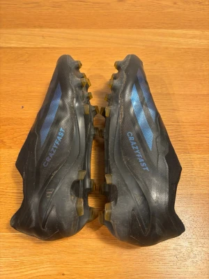 Adidas Crazyfast svarta fotbollsskor - Säljer ett par svarta Adidas Crazyfast fotbollsskor med blå detaljer och klassiska tre ränder på sidan. Skorna har dobbar för bra grepp på planen och är tillverkade i syntetmaterial för lätt känsla och snabbhet. Perfekta för dig som vill ha stil och fart på fotbollsplanen. En nackdel är hål i högra tån men är spelbara.