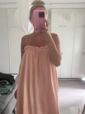 Ljusrosa långklänning med smala axelband - Säljer en ljusrosa långklänning med smala axelband och rynkad överdel. Klänningen har en luftig och rak passform, perfekt för varma dagar. Materialet känns lätt och bekvämt mot huden. Enkel och stilren design som ger en avslappnad vibe.